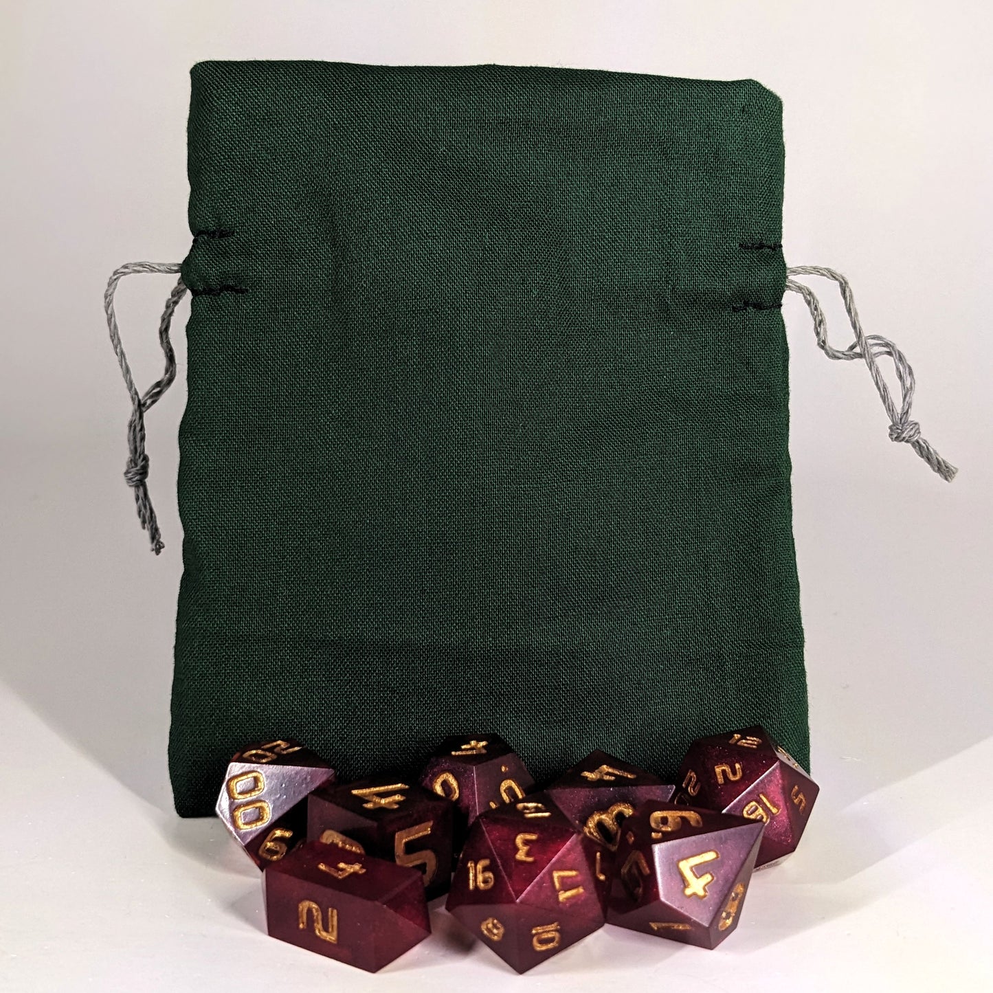 A small, handmade dice bag that is dark green stands upright on a white background with a set of red dnd dice with gold numbers sitting in front of it.