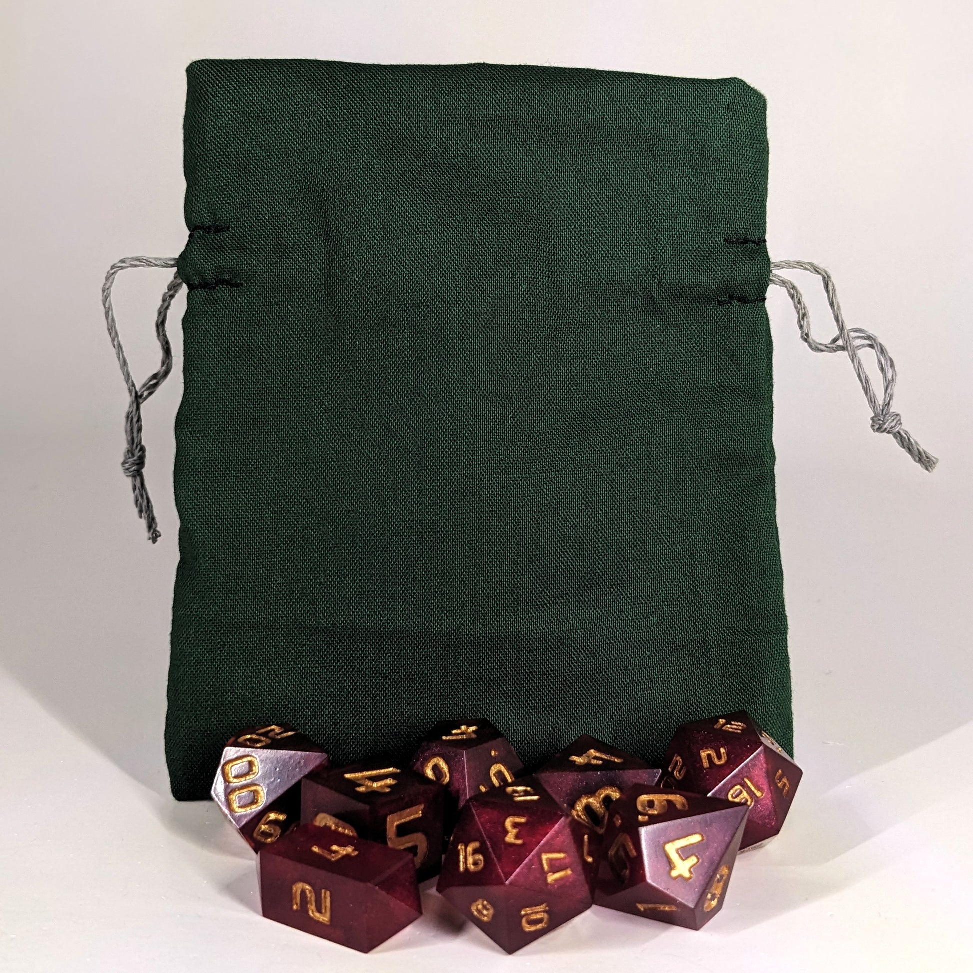 A small, handmade dice bag that is dark green stands upright on a white background with a set of red dnd dice with gold numbers sitting in front of it.