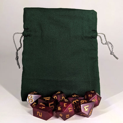 A small, handmade dice bag that is dark green stands upright on a white background with a set of red dnd dice with gold numbers sitting in front of it.