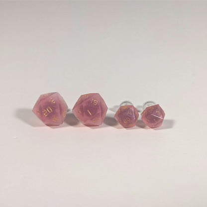 Two pairs of handmade d20 earrings with titanium hardware that are bright pink with gold numbers on a white background sit in a line with the larger pair on the left and the smaller pair on the right. The earrings on the left of each pair centers the 20 face and the earrings on the right of each pair centers the 1 face.