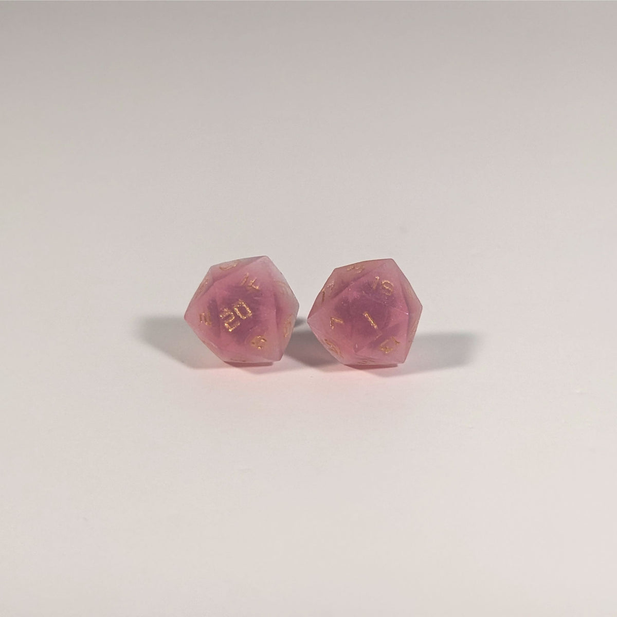 A single pair of the larger size handmade d20 earrings with titanium hardware that are bright pink with gold numbers on a white background. The earring on the left centers the 20 face and the earring on the right centers the 1 face.