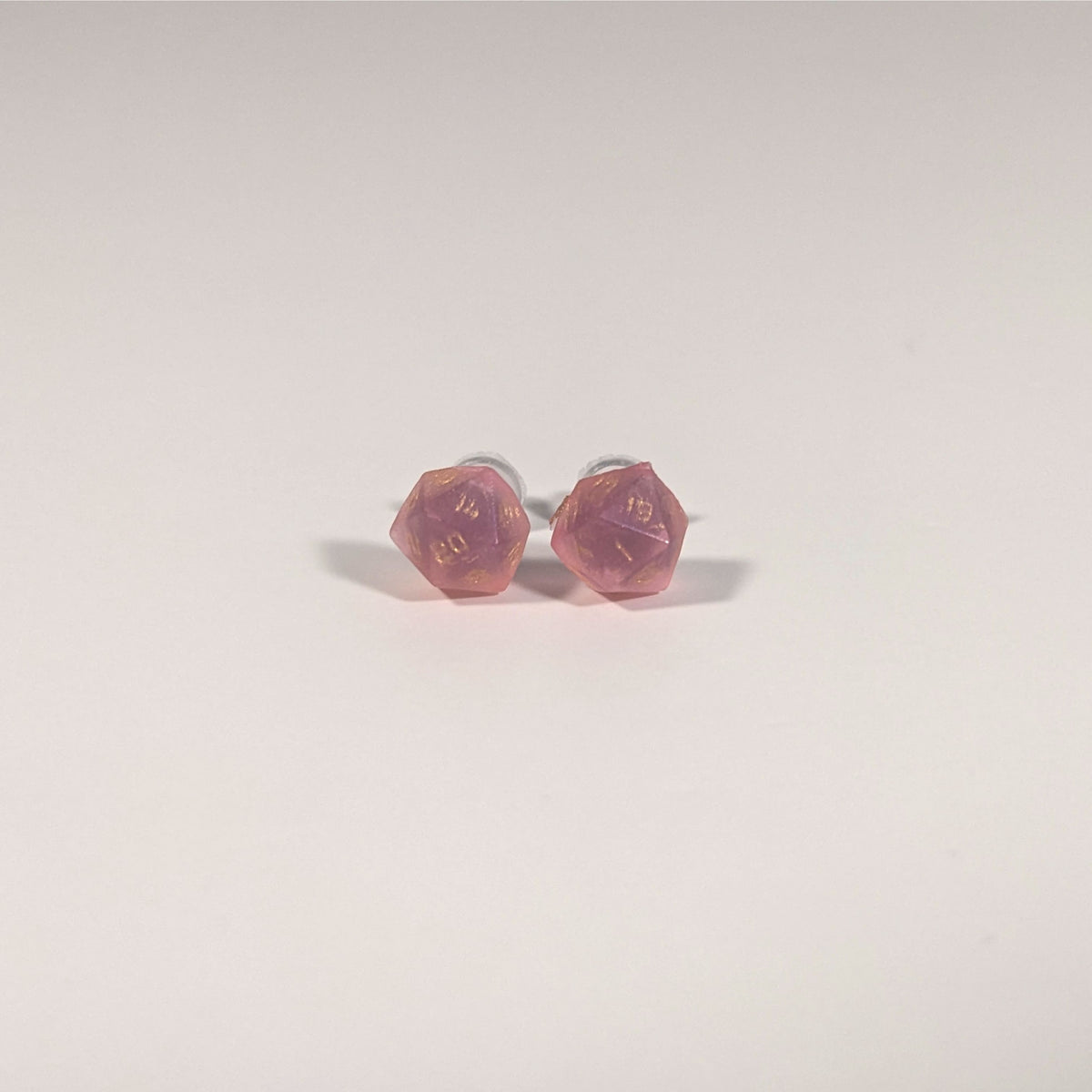 A single pair of the smaller size handmade d20 earrings with titanium hardware that are bright pink with gold numbers on a white background. The earring on the left centers the 20 face and the earring on the right centers the 1 face.