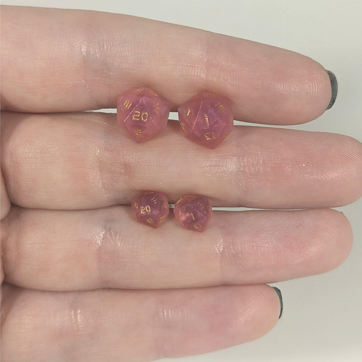 Two pairs of handmade d20 earrings with titanium hardware that are bright pink with gold numbers held between the fingers of a flat hand. The larger pair is between the pointer and middle finger, the smaller between the middle and ring finger. The earrings on the left of each pair centers the 20 face and the earrings on the right of each pair centers the 1 face.