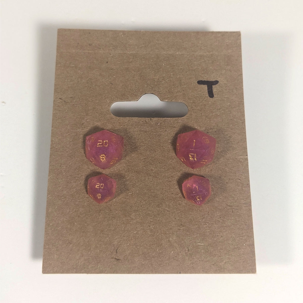 Two pairs of handmade d20 earrings with titanium hardware that are bright pink with gold numbers on a kraft brown earring card with a T in the top right corner. The earrings on the left of each pair centers the 20 face and the earrings on the right of each pair centers the 1 face.