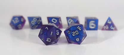 A straight on photo of a standard set of DnD dice that are blue and purple with gray numbers focusing on 2 d20s in the center. The d20 on the left has the 1 face centered and the d20 on the right has the 20 face centered. The other 6 dice are lined up in the background but are out of focus, while the d20s are in sharp focus.