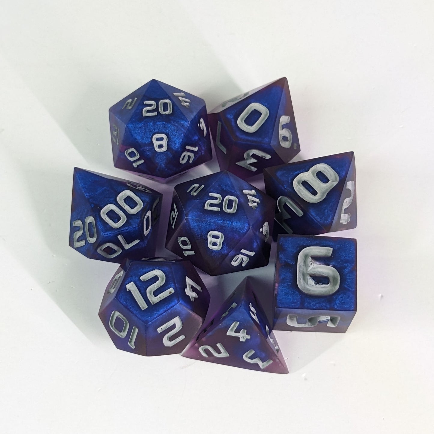  A complete set of 8 handmade artisan dice that are blue and purple with grey numbers. The DnD dice set is arranged in a circle and features 2 D20s, one of which is in the center surrounded by the other 7 dice. 