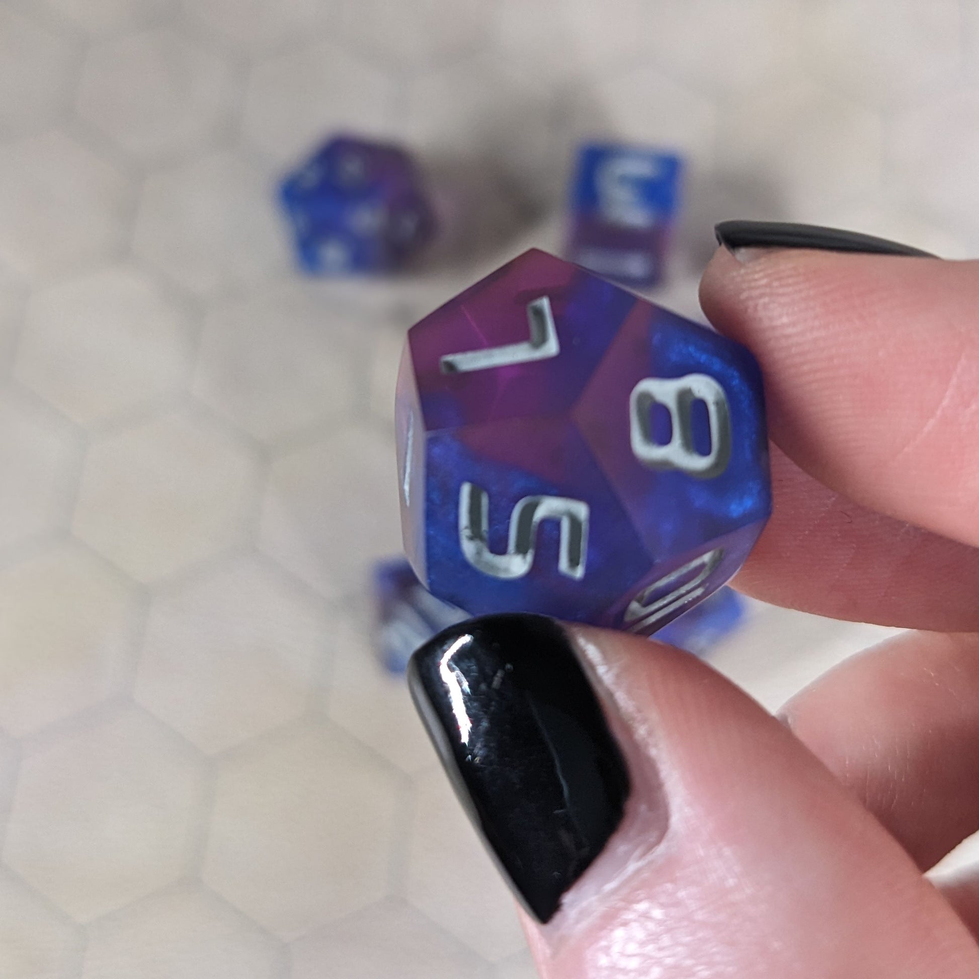 A d12 that is blue and purple with gray numbers is held up to the camera to show off the color swirls. The 7, 5, and 8 faces are angled toward the camera.