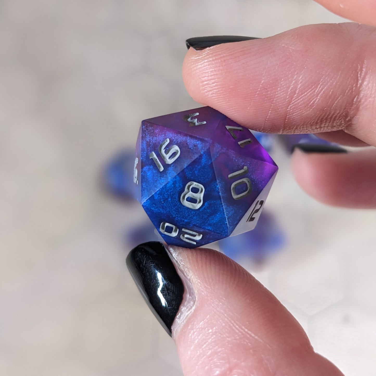 A d20 that is blue and purple with gray numbers is held up to the camera to show off the color swirls. The 8 face is angled toward the camera.