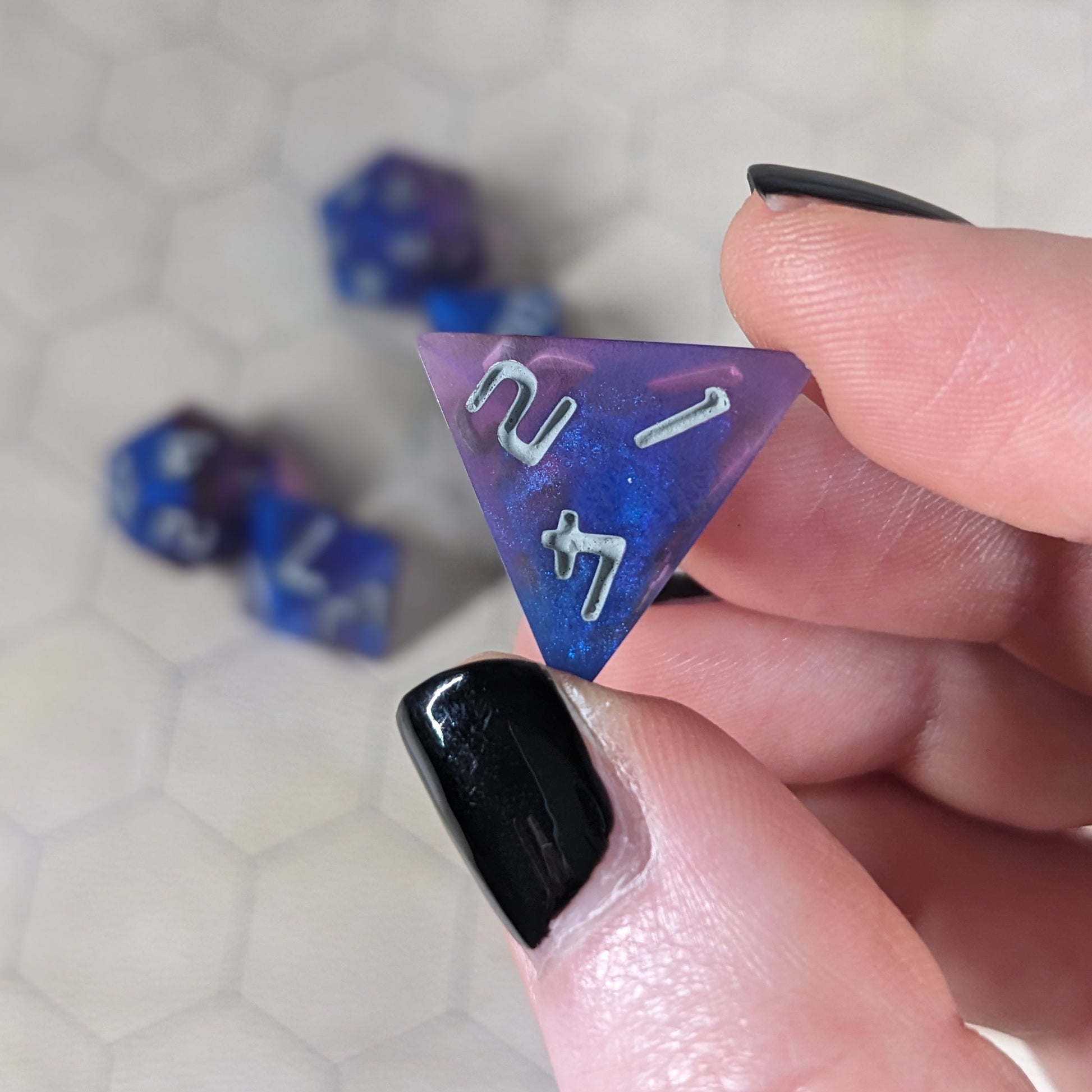 A d4 that is blue and purple with gray numbers is held up to the camera to show off the color swirls. The 1, 2, 4 face is angled toward the camera.