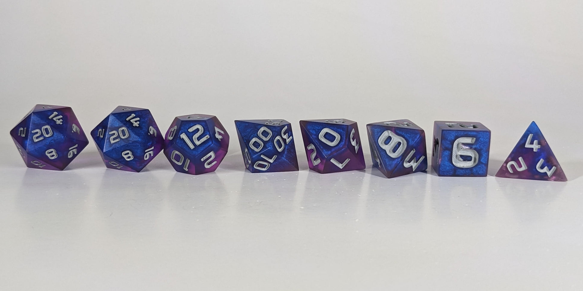 A straight on photo of a complete set of polyhedral dice that are blue and purple with gray numbers arranged in a straight line. They are lined up with the 2 d20s on the left going down to the d4 on the right side. The highest value number of each die is facing forward.