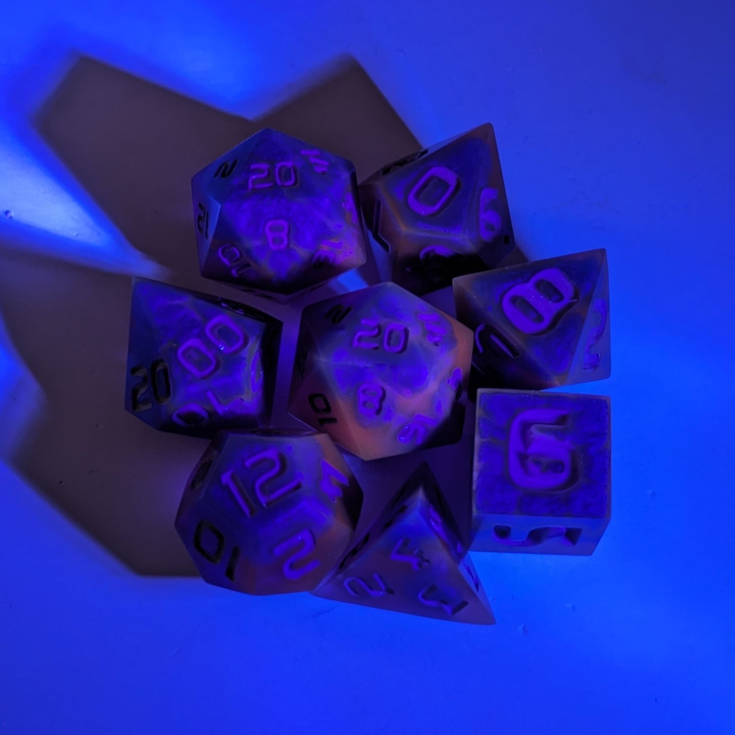  A complete set of 8 handmade artisan dice that are blue and purple with grey numbers under a UV light. The DnD dice set is arranged in a circle and features 2 D20s, one of which is in the center surrounded by the other 7 dice.  The purple colorant glows pinkish under the UV light, and the background glows blue from the light.