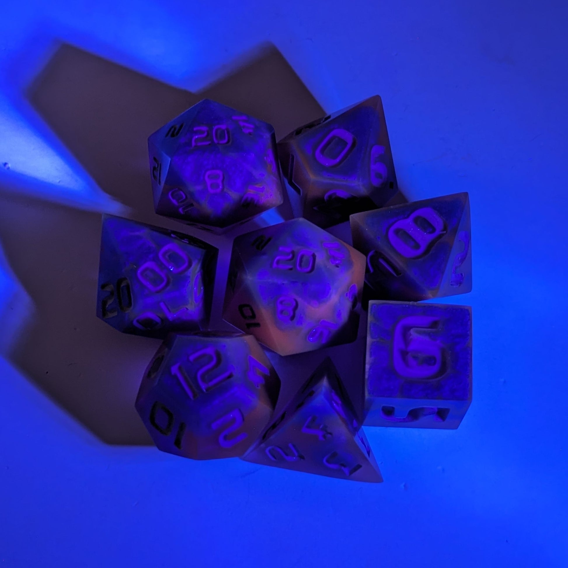  A complete set of 8 handmade artisan dice that are blue and purple with grey numbers under a UV light. The DnD dice set is arranged in a circle and features 2 D20s, one of which is in the center surrounded by the other 7 dice.  The purple colorant glows pinkish under the UV light, and the background glows blue from the light.