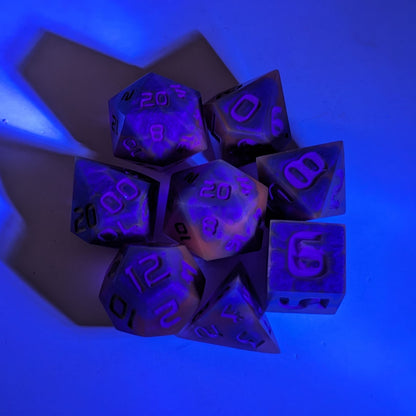  A complete set of 8 handmade artisan dice that are blue and purple with grey numbers under a UV light. The DnD dice set is arranged in a circle and features 2 D20s, one of which is in the center surrounded by the other 7 dice.  The purple colorant glows pinkish under the UV light, and the background glows blue from the light.
