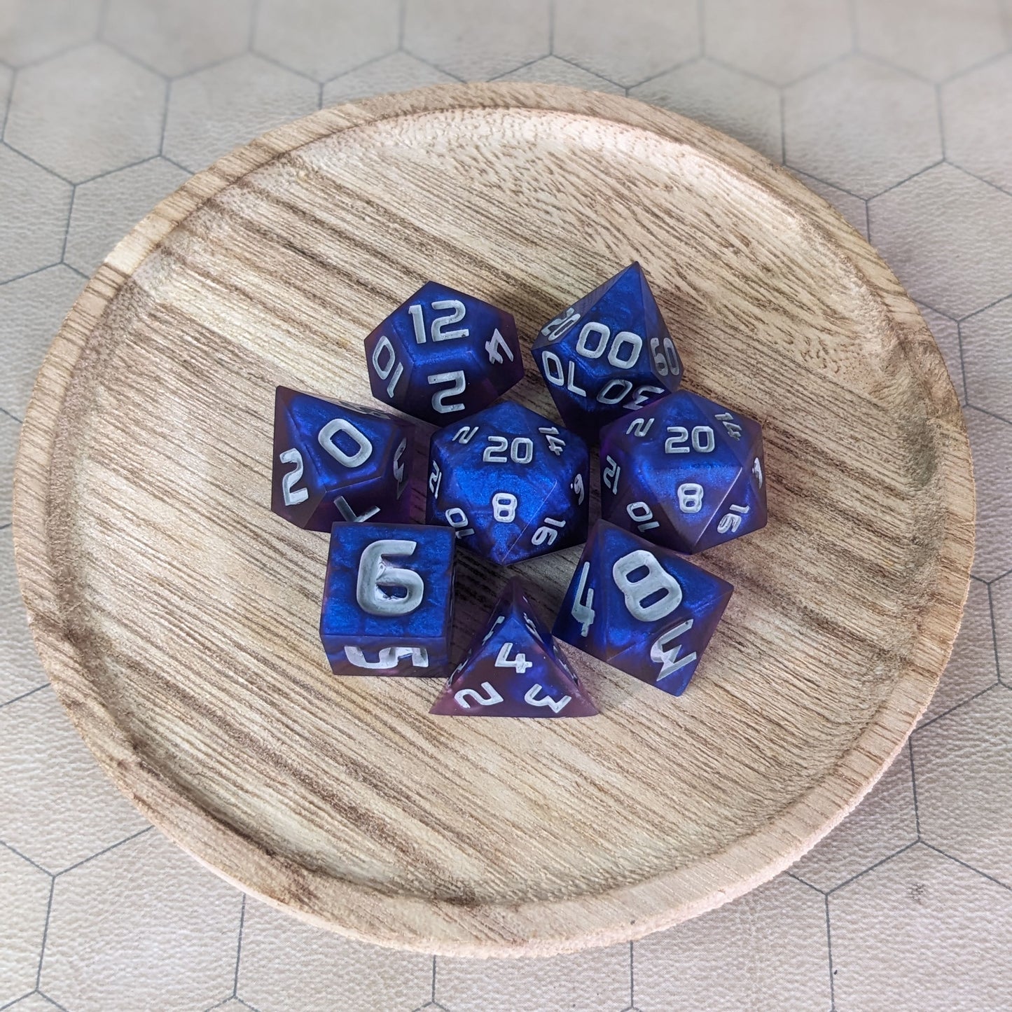  A complete set of 8 handmade artisan dice that are blue and purple with grey numbers. The DnD dice set is on a wooded circle and the dice are arranged in a circle and features 2 D20s, one of which is in the center surrounded by the other 7 dice. 