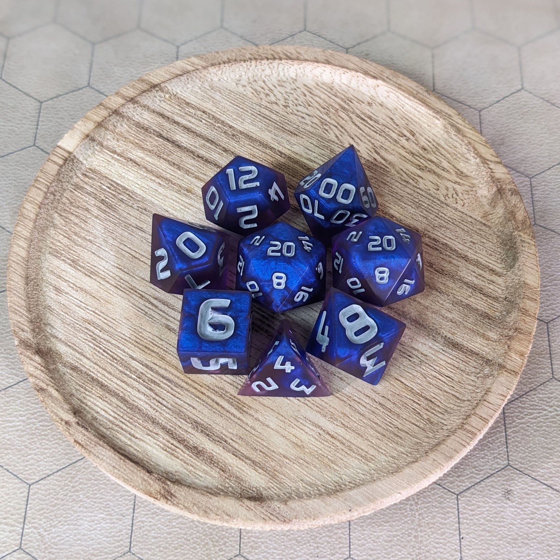  A complete set of 8 handmade artisan dice that are blue and purple with grey numbers. The DnD dice set is on a wooded circle and the dice are arranged in a circle and features 2 D20s, one of which is in the center surrounded by the other 7 dice. 