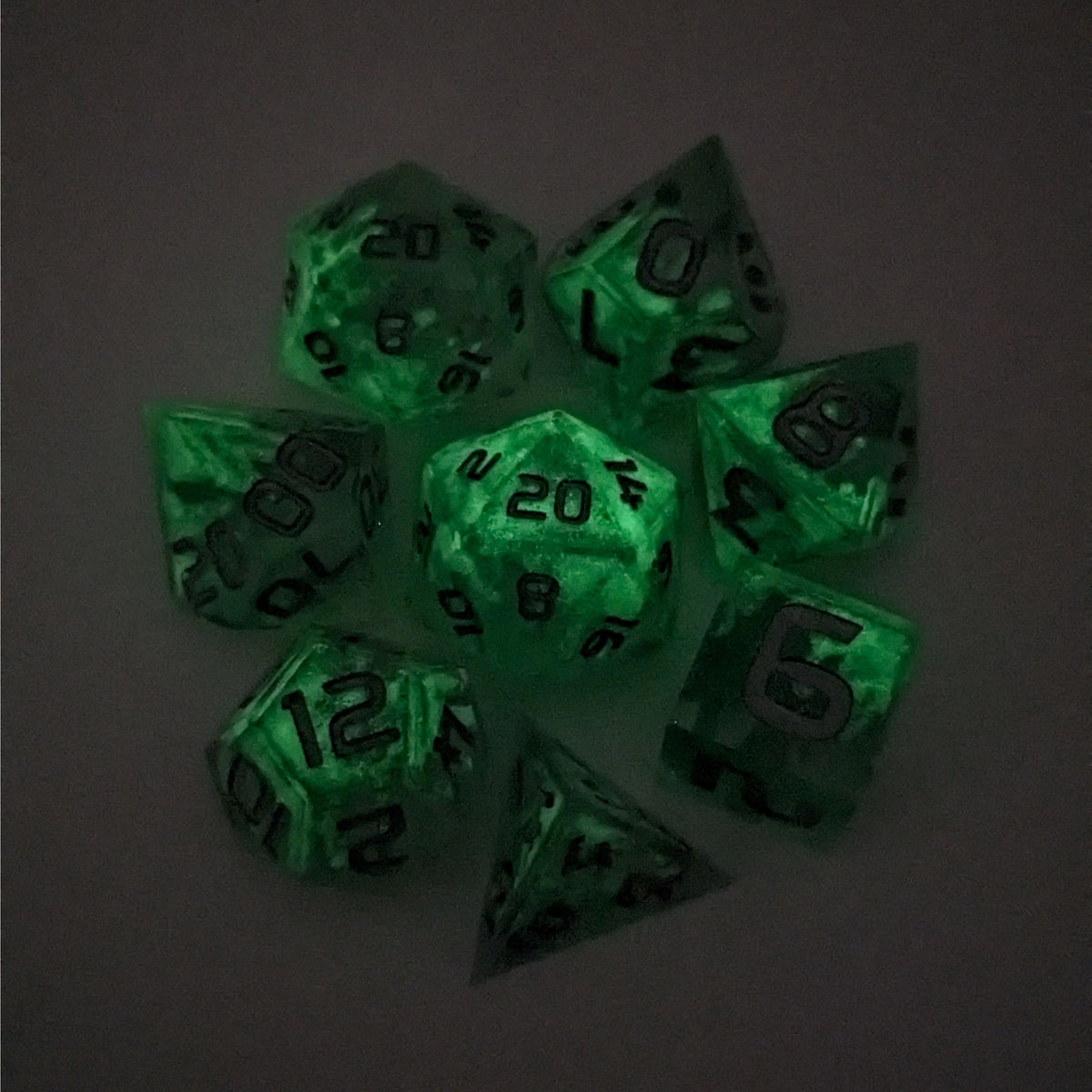 A complete set of 8 handmade artisan dice that are clear with a hand painted green glow in the dark ooze design and silver numbers. The DnD dice set is arranged in a circle and features 2 D20s, one of which is in the center surrounded by the other 7 dice on a white background. This photo was taken in the dark to show the ooze design glowing in the dark.