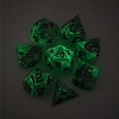A complete set of 8 handmade artisan dice that are clear with a hand painted green glow in the dark ooze design and silver numbers. The DnD dice set is arranged in a circle and features 2 D20s, one of which is in the center surrounded by the other 7 dice on a white background. This photo was taken in the dark to show the ooze design glowing in the dark.