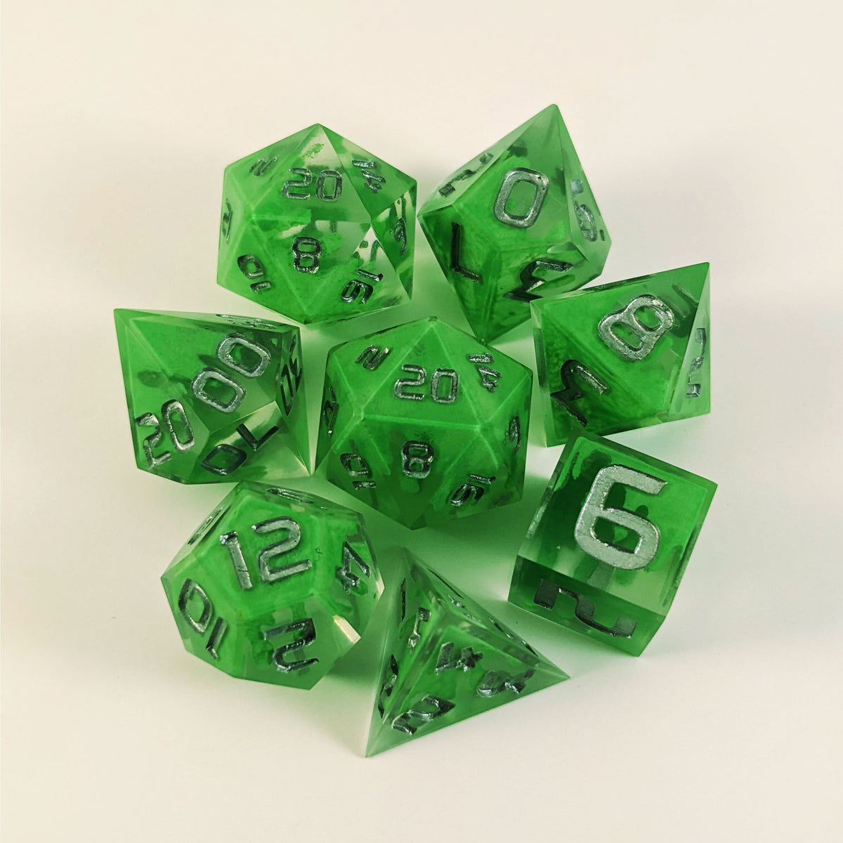 A complete set of 8 handmade artisan dice that are clear with a hand painted green glow in the dark ooze design and silver numbers. The DnD dice set is arranged in a circle and features 2 D20s, one of which is in the center surrounded by the other 7 dice on a white background.