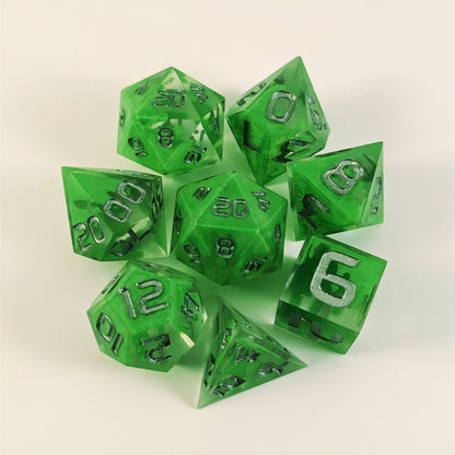 A complete set of 8 handmade artisan dice that are clear with a hand painted green glow in the dark ooze design and silver numbers. The DnD dice set is arranged in a circle and features 2 D20s, one of which is in the center surrounded by the other 7 dice on a white background.