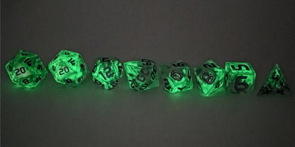 A straight on photo of a complete set of polyhedral dice that are clear with a hand painted green glow in the dark ooze design and silver numbers arranged in a straight line. They are lined up with the 2 d20s on the left going down to the d4 on the right side. The highest value number of each die is facing forward. This photo is taken in the dark to show the ooze design glowing green in the dark.