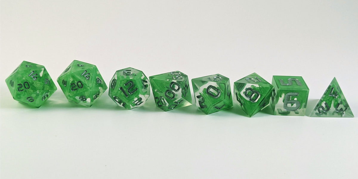 A straight on photo of a complete set of polyhedral dice that are clear with a hand painted green glow in the dark ooze design and silver numbers arranged in a straight line. They are lined up with the 2 d20s on the left going down to the d4 on the right side. The highest value number of each die is facing forward.
