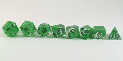 A straight on photo of a complete set of polyhedral dice that are clear with a hand painted green glow in the dark ooze design and silver numbers arranged in a straight line. They are lined up with the 2 d20s on the left going down to the d4 on the right side. The highest value number of each die is facing forward.