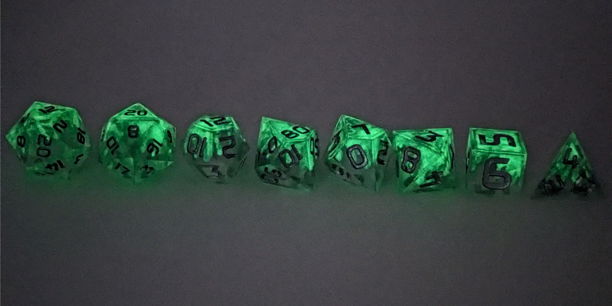 A straight on photo of a complete set of polyhedral dice that are clear with a hand painted green glow in the dark ooze design and silver numbers arranged in a straight line. They are lined up with the 2 d20s on the left going down to the d4 on the right side. The dice are arranged so the ooze design is dripping downward. This photo is taken in the dark to show the ooze design glowing green in the dark.