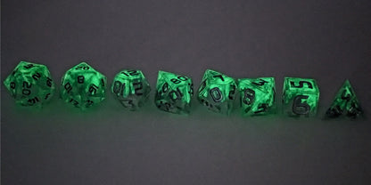 A straight on photo of a complete set of polyhedral dice that are clear with a hand painted green glow in the dark ooze design and silver numbers arranged in a straight line. They are lined up with the 2 d20s on the left going down to the d4 on the right side. The dice are arranged so the ooze design is dripping downward. This photo is taken in the dark to show the ooze design glowing green in the dark.