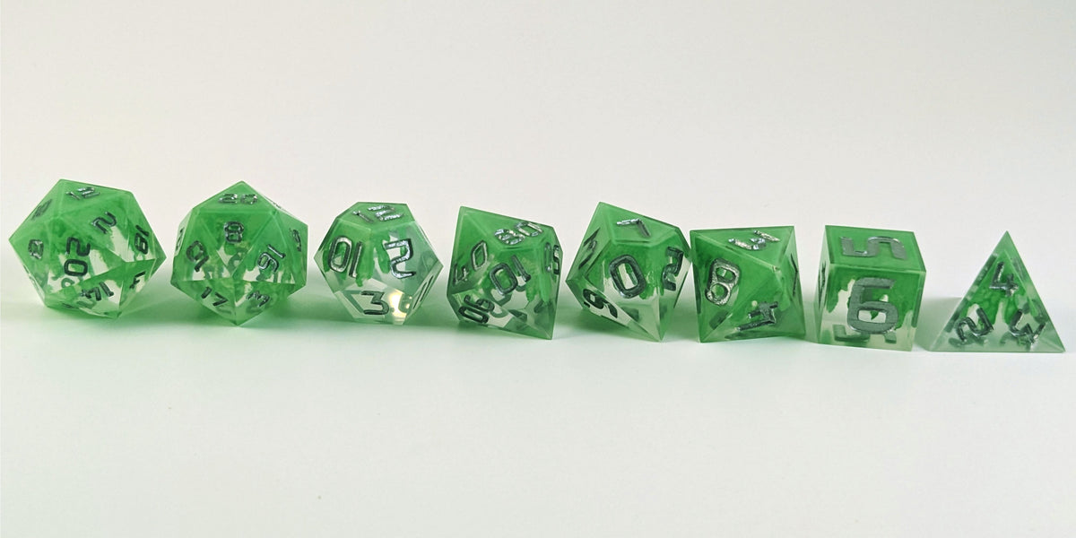 A straight on photo of a complete set of polyhedral dice that are clear with a hand painted green glow in the dark ooze design and silver numbers arranged in a straight line. They are lined up with the 2 d20s on the left going down to the d4 on the right side. The dice are arranged so the ooze design is dripping downward.