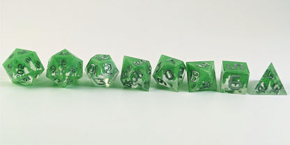 A straight on photo of a complete set of polyhedral dice that are clear with a hand painted green glow in the dark ooze design and silver numbers arranged in a straight line. They are lined up with the 2 d20s on the left going down to the d4 on the right side. The dice are arranged so the ooze design is dripping downward.
