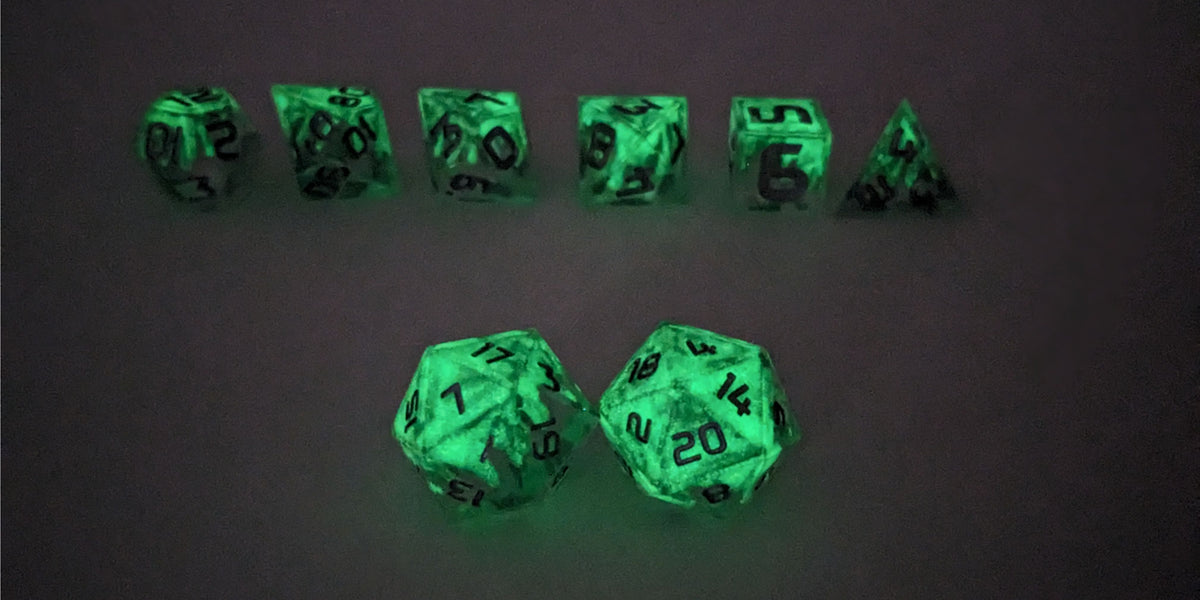 A straight on photo of a standard set of DnD dice that are clear with a hand painted green glow in the dark ooze design and silver numbers focusing on 2 d20s in the center. The d20 on the left has the 1 face centered and the d20 on the right has the 20 face centered. The other 6 dice are lined up in the background but are out of focus, while the d20s are in sharp focus. This photo is taken in the dark to show the ooze design glowing green in the dark.