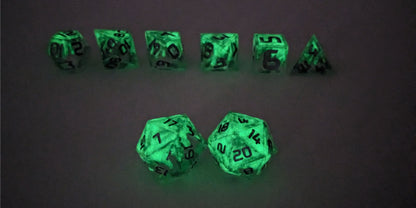 A straight on photo of a standard set of DnD dice that are clear with a hand painted green glow in the dark ooze design and silver numbers focusing on 2 d20s in the center. The d20 on the left has the 1 face centered and the d20 on the right has the 20 face centered. The other 6 dice are lined up in the background but are out of focus, while the d20s are in sharp focus. This photo is taken in the dark to show the ooze design glowing green in the dark.