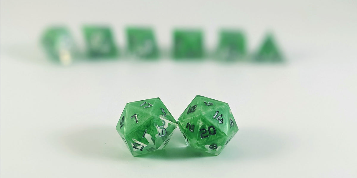A straight on photo of a standard set of DnD dice that are clear with a hand painted green glow in the dark ooze design and silver numbers focusing on 2 d20s in the center. The d20 on the left has the 1 face centered and the d20 on the right has the 20 face centered. The other 6 dice are lined up in the background but are out of focus, while the d20s are in sharp focus.