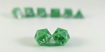 A straight on photo of a standard set of DnD dice that are clear with a hand painted green glow in the dark ooze design and silver numbers focusing on 2 d20s in the center. The d20 on the left has the 1 face centered and the d20 on the right has the 20 face centered. The other 6 dice are lined up in the background but are out of focus, while the d20s are in sharp focus.