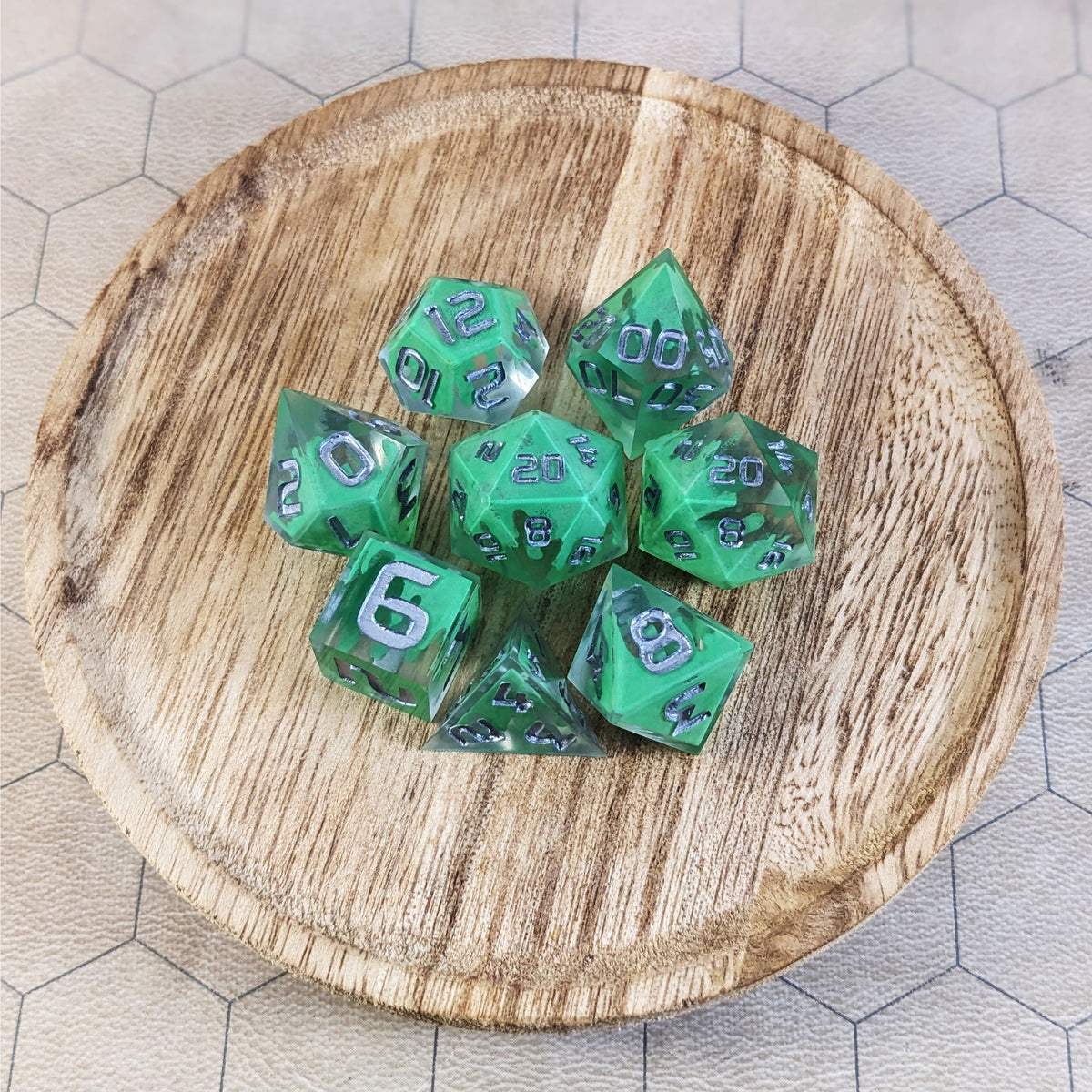 A complete set of 8 handmade artisan dice that are clear with a hand painted green glow in the dark ooze design and silver numbers. The DnD dice set is on a wooded circle and the dice are arranged in a circle and features 2 D20s, one of which is in the center surrounded by the other 7 dice.