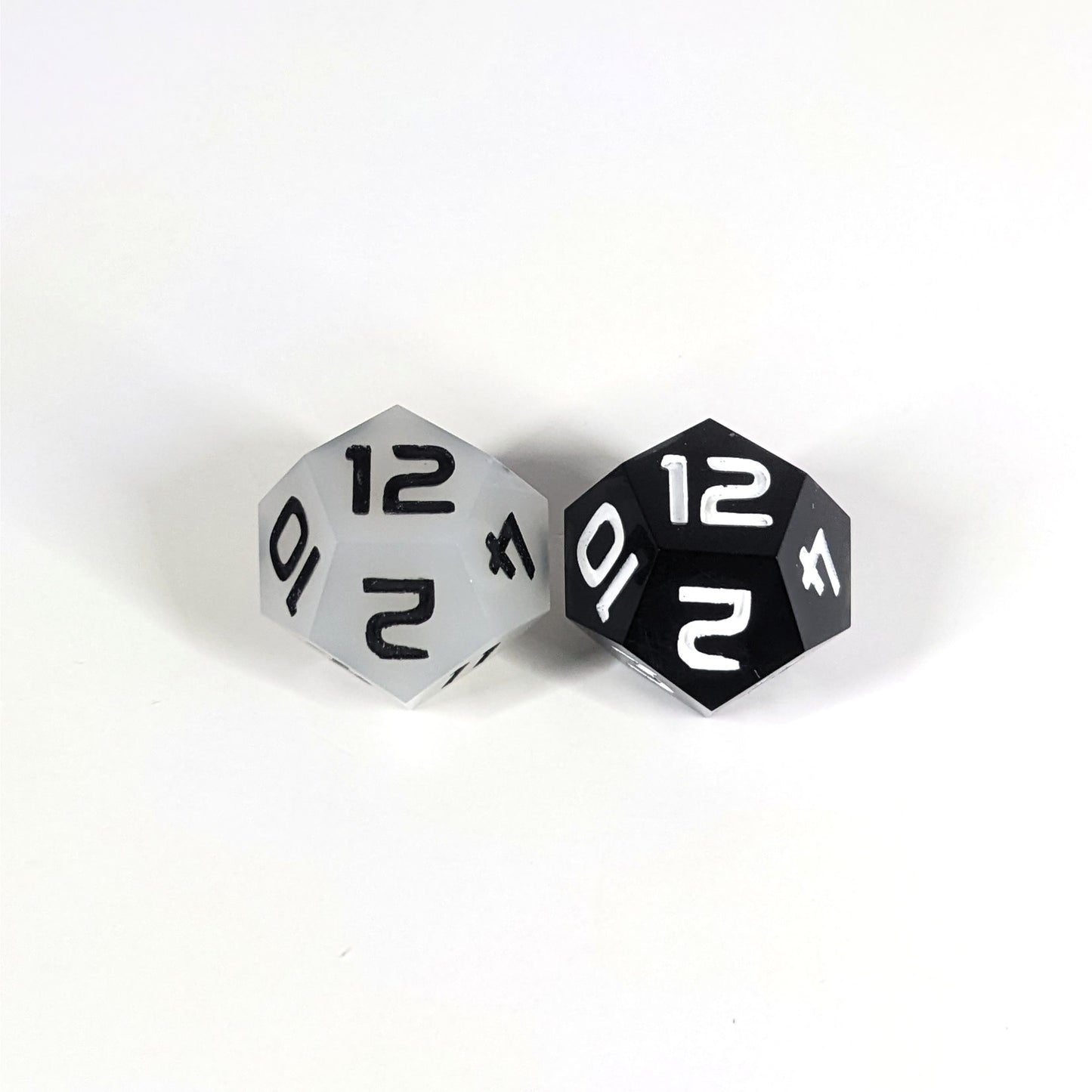 Two handmade d12s sit with the 12 faces up on a white background. The die on the left is white with black numbers, the die on the right is black with white numbers.