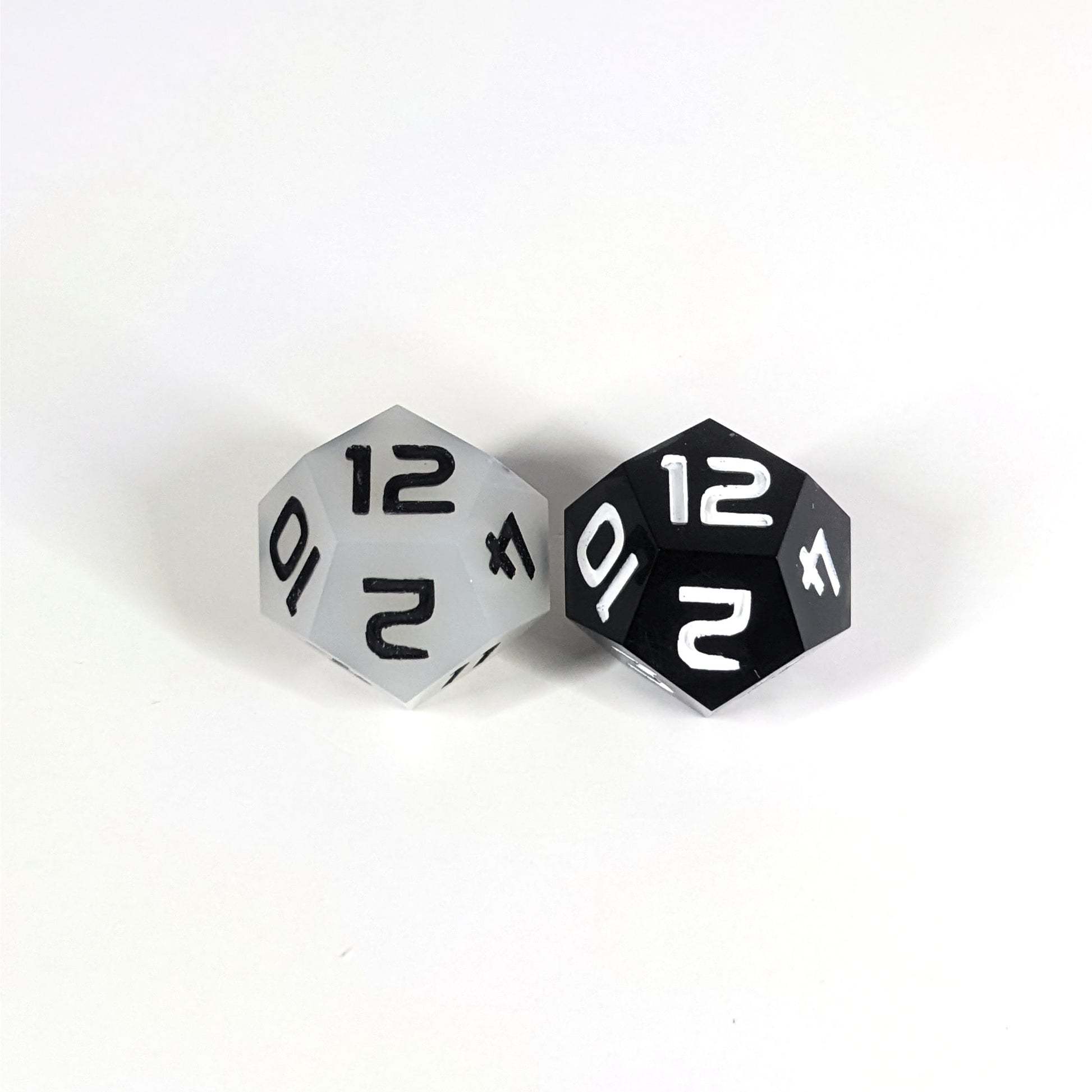 Two handmade d12s sit with the 12 faces up on a white background. The die on the left is white with black numbers, the die on the right is black with white numbers.