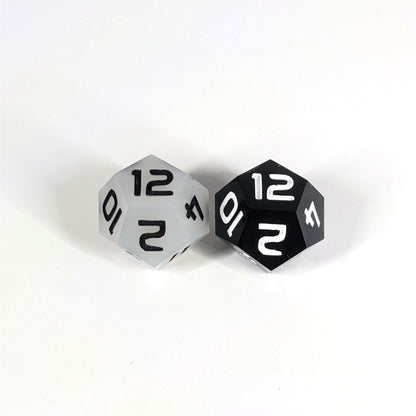 Two handmade d12s sit with the 12 faces up on a white background. The die on the left is white with black numbers, the die on the right is black with white numbers.
