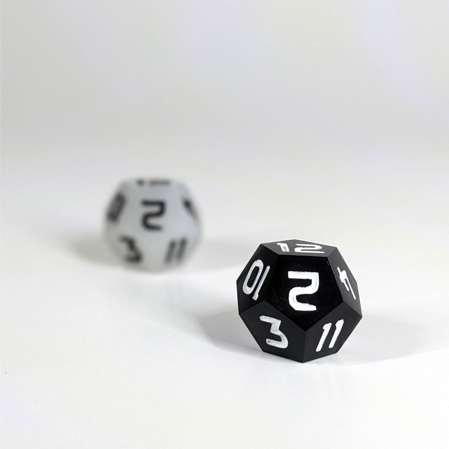 A low angle photo of two d12s. The white d12  with black numbers is on the left and is further from the camera and out of focus. The black d12 with white numbers is on the right and is closer to the camera and in sharp focus.