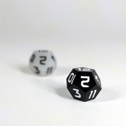A low angle photo of two d12s. The white d12  with black numbers is on the left and is further from the camera and out of focus. The black d12 with white numbers is on the right and is closer to the camera and in sharp focus.