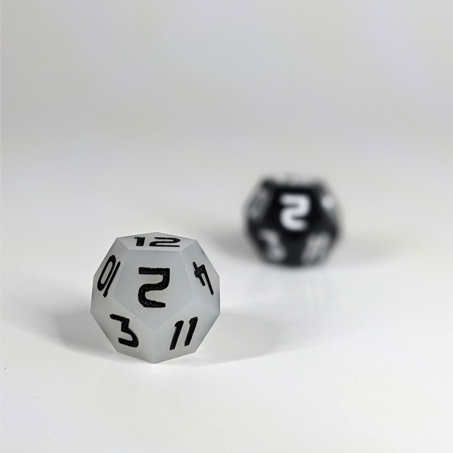 A low angle photo of two d12s. The white d12 with black numbers is on the left and is closer to the camera in sharp focus. The black d12 with white numbers is on the right and is further from the camera and out of focus.