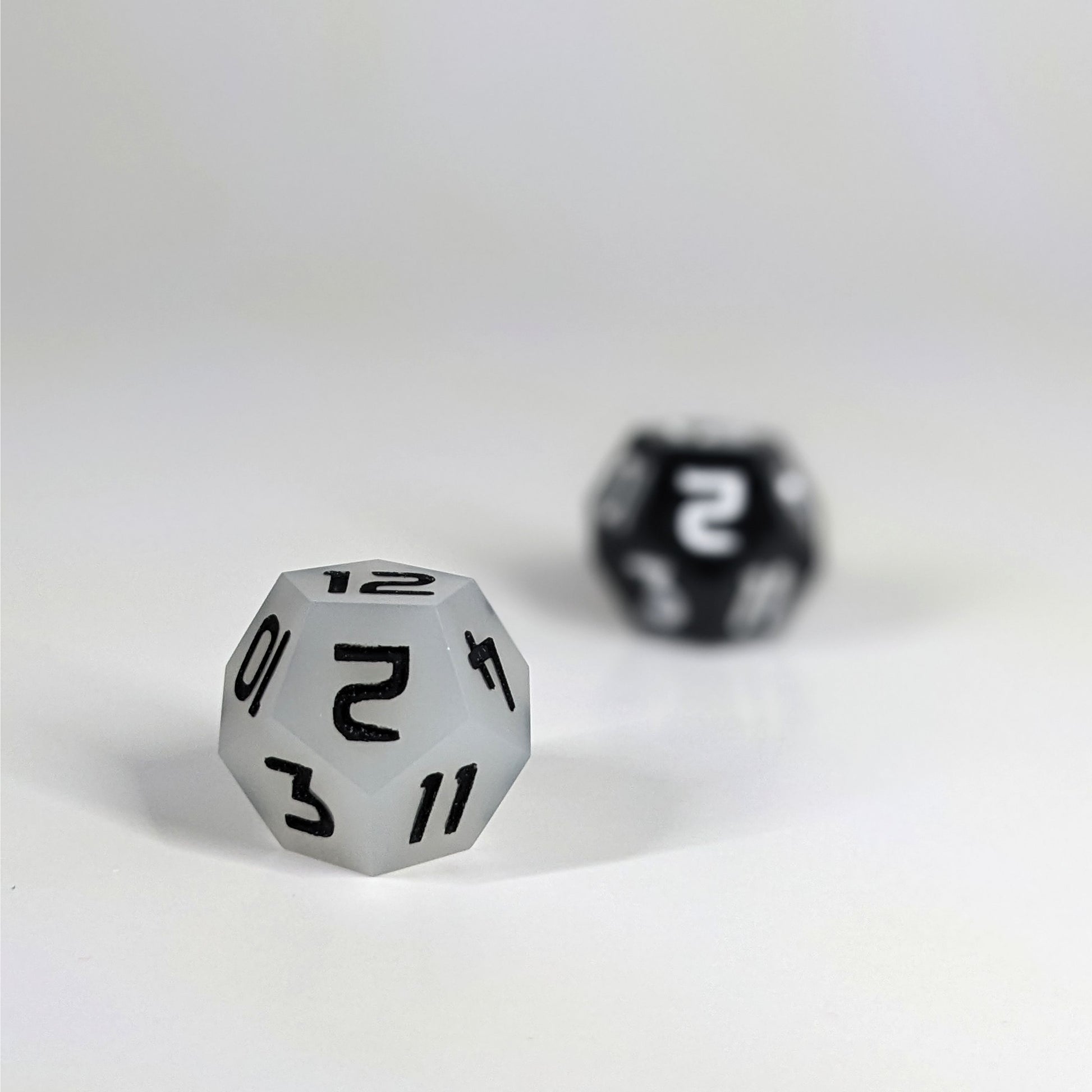 A low angle photo of two d12s. The white d12 with black numbers is on the left and is closer to the camera in sharp focus. The black d12 with white numbers is on the right and is further from the camera and out of focus.