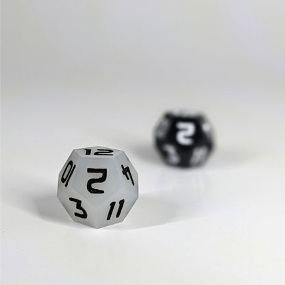 A low angle photo of two d12s. The white d12 with black numbers is on the left and is closer to the camera in sharp focus. The black d12 with white numbers is on the right and is further from the camera and out of focus.