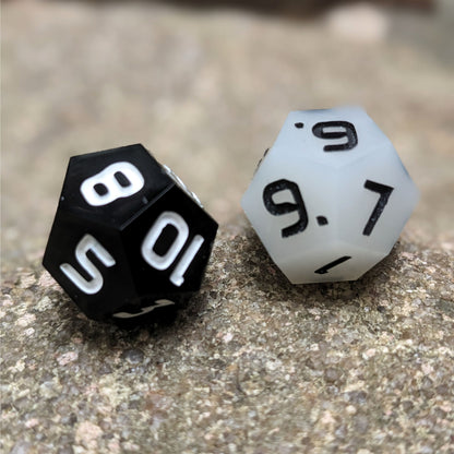 Two handmade d12s sit on a large rock. One die is white with black numbers and the other is black with white numbers.