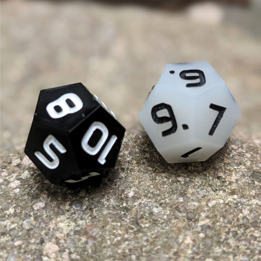 Two handmade d12s sit on a large rock. One die is white with black numbers and the other is black with white numbers.