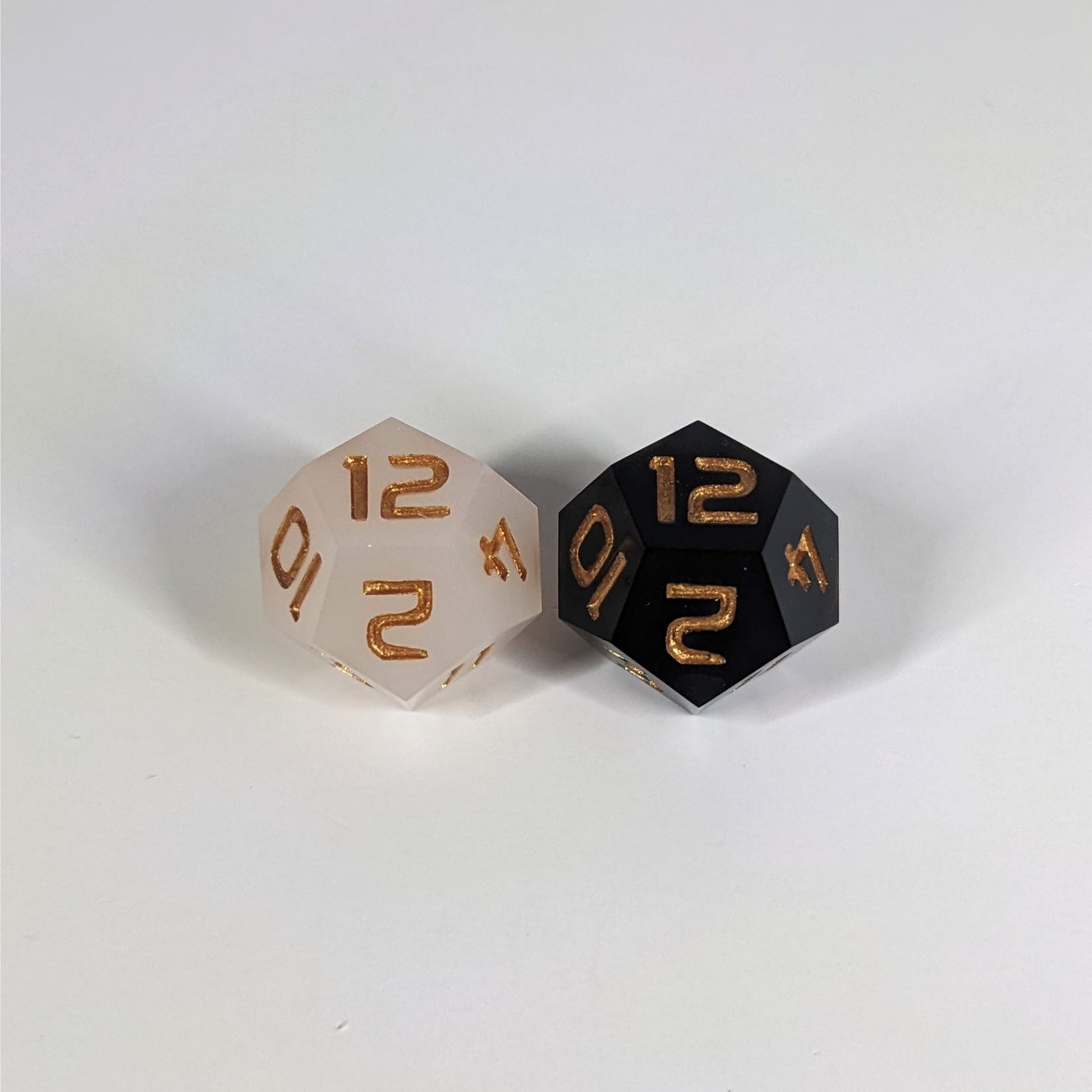 Two handmade d12s sit with the 12 faces up on a white background. The die on the left is white, the die on the right is black, and both dice have gold numbers.