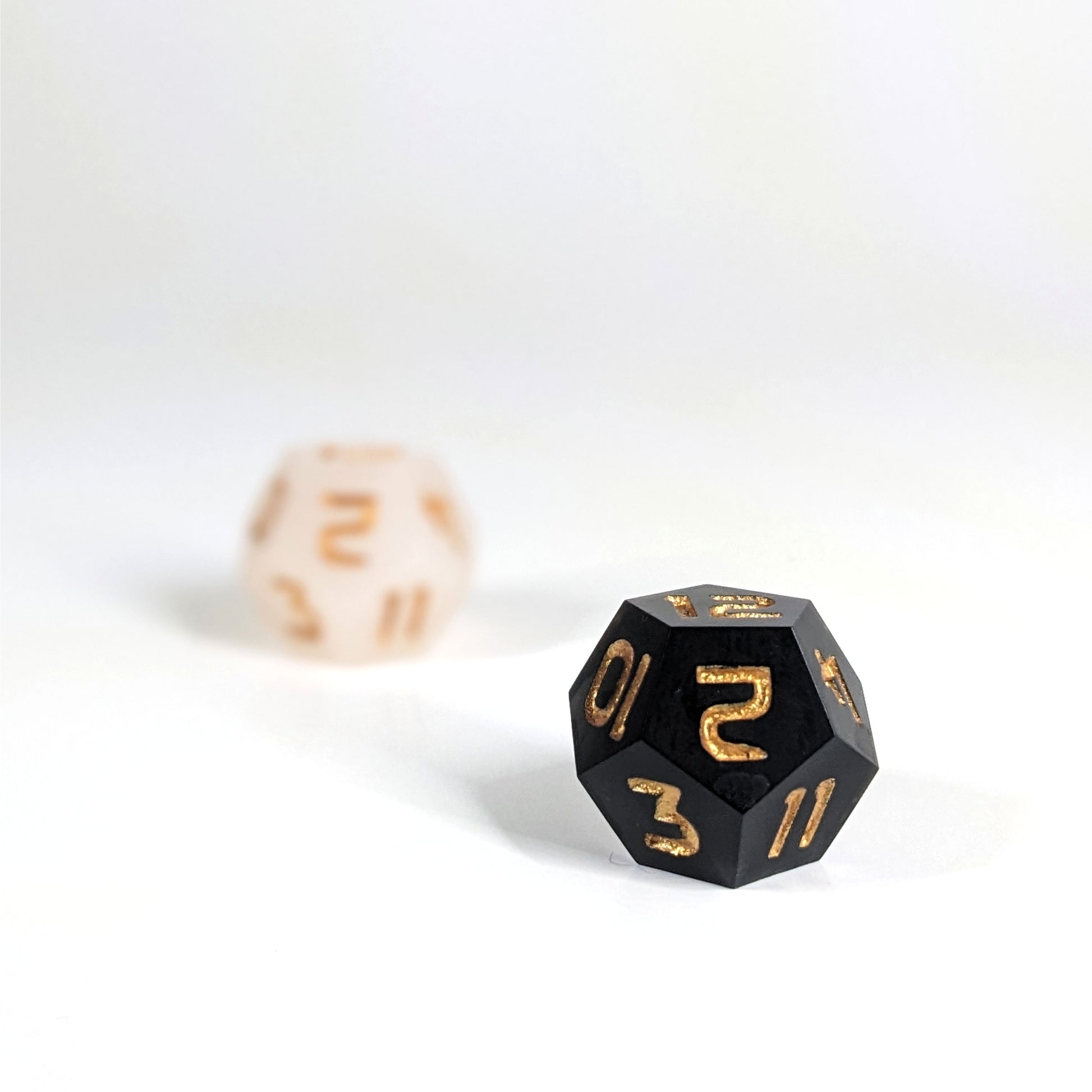 A low angle photo of two d12s. The white d12 is on the left and is further from the camera and out of focus. The black d12 is on the right and is closer to the camera and in sharp focus. Both dice have gold numbers.