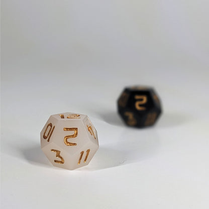 A low angle photo of two d12s. The white d12 is on the left and is closer to the camera in sharp focus. The black d12 is on the right and is further from the camera and out of focus. Both dice have gold numbers.