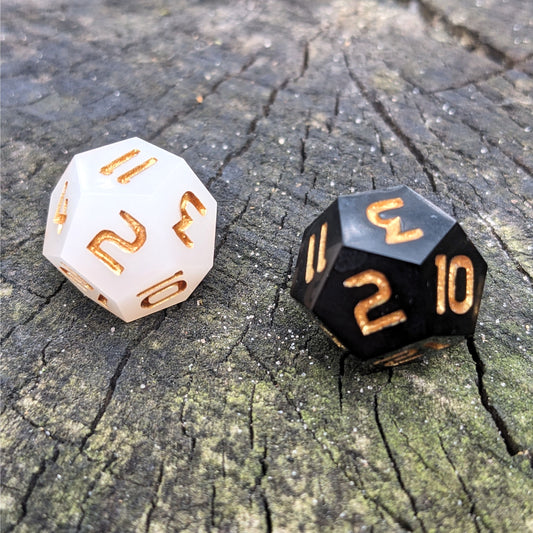 Two handmade d12s sit on a wood stump. One die is white and the other is black, both dice have gold numbers.