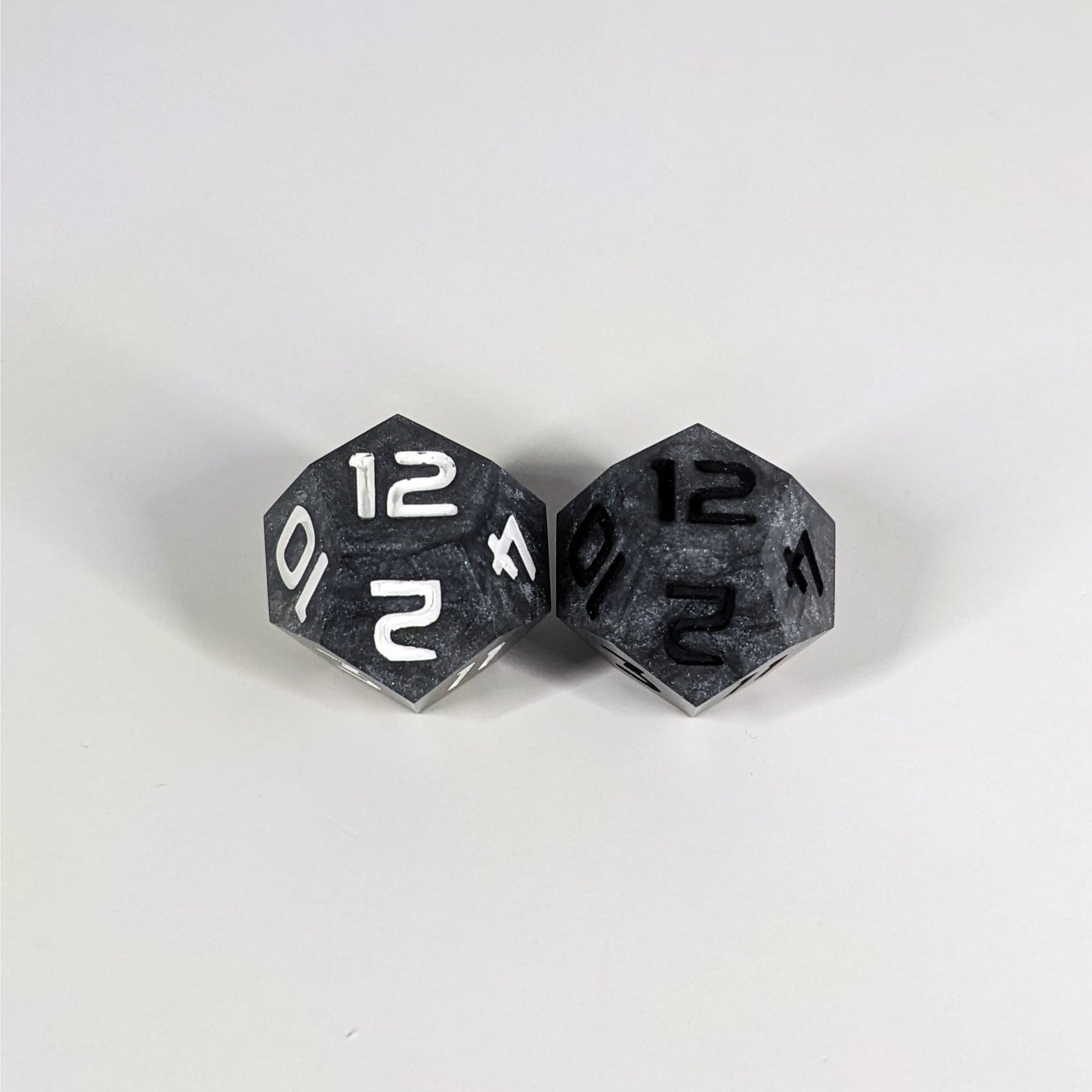 Two handmade d12s sit with the 12 faces up on a white background. The dice are dark silver, and while they also have a touch of gold the gold is not visible at this angle. The die on the left has white numbers, the die on the right has black numbers.